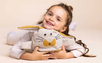 Glückliches Mädchen liegt mit Kuscheltier in Hasenform auf hellem Untergrund. Das Bild vermittelt Wärme, Natürlichkeit und Nachhaltigkeit als Kernwerte für umweltfreundliche Möbel und gesundes Wohnen.